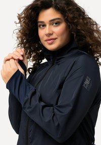 Granatowa lekka kurtka z podwyższonym kołnierzem, zamkiem z przodu, teksturowanymi panelami na ramionach oraz akcentami z logo na rękawie.