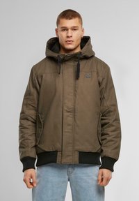 Geacă căptușită verde măsliniu cu glugă, cu accente negre, manșete elastice și un patch cu logo pe piept. Textură netedă.
