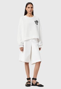 Felpa bianca corta con logo grafico nero, abbinata a pantaloni corti bianchi a gamba larga. Sandali neri con cinturini alla caviglia completano il look.