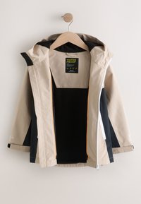 Veste imperméable beige et noire avec capuche, suspendue ouverte sur un cintre en bois, montrant une doublure polaire noire et une étiquette à l'intérieur du col.