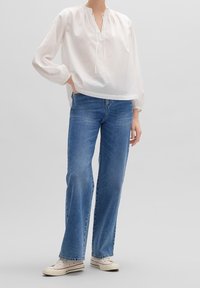 Personne portant une blouse blanche à manches longues ample, un jean droit bleu et des baskets en toile beige sur fond clair.