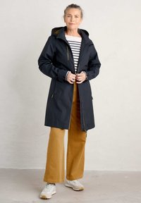 Giacca impermeabile blu navy con cappuccio, zip frontale e tasche laterali, abbinata a pantaloni larghi color senape e sneakers bianche. Sotto una maglietta a righe.