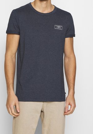 Mann trägt ein dunkelgraues T-Shirt mit Rundhalsausschnitt und einem kleinen weißen Aufnäher auf der linken Brust sowie beige Hosen, steht vor einem einfarbigen Hintergrund.