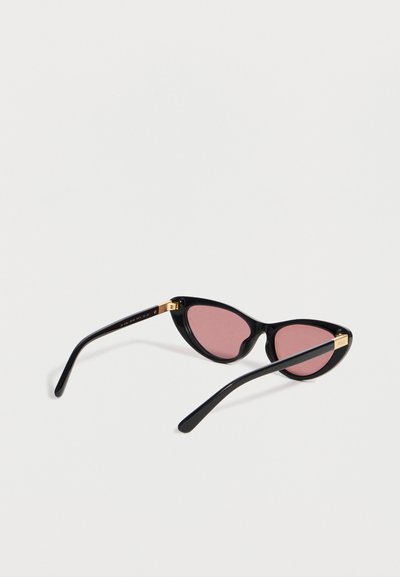 Μαύρα γυαλιά ηλίου cat-eye με ροζ φακούς και χρυσές λεπτομέρειες στα μπράτσα, τοποθετημένα σε λευκή επιφάνεια.
