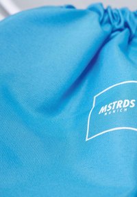 Blå snørepose laget av teksturert stoff, med en hvit logo som har "MSTRDS MUNICH" på overflaten.
