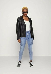 Jaqueta de motoqueiro em couro preto, top branco, jeans de retalhos azul claro e tênis pretos com solas brancas. Modelo usando óculos de sol.