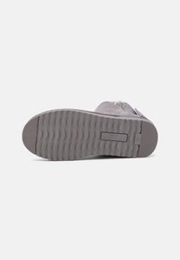 Chaussure grise à enfiler avec une semelle en caoutchouc texturée à motif ondulé, matière supérieure douce et une sangle latérale pour le réglage.