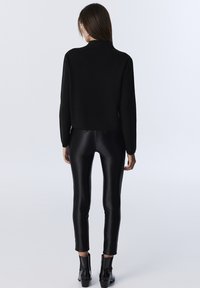 Žena stojící zády, oblečená v černém dlouhém rukávu, lesklých černých legínách a černých kotníkových botách, na hladkém světlém pozadí.