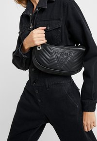 Borsa nera trapuntata con motivo a spina di pesce, dettagli in argento e logo "GUESS". Indossata a tracolla su un outfit nero.