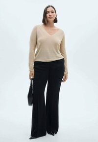 Beige Langarm-V-Ausschnitt-Oberteil kombiniert mit hochgeschnittenen schwarzen weiten Hosen. Das Model hält eine schwarze Handtasche und trägt spitze schwarze Schuhe.