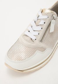 Gros plan d'une basket beige avec des lacets blancs, une fermeture éclair latérale, un panneau avant texturé et une semelle blanche avec une bordure beige.