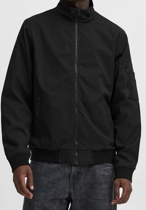 Mann trägt eine schlichte schwarze Bomberjacke mit Reißverschluss, gerippten Bündchen und einer Reißverschlusstasche am linken Ärmel, kombiniert mit dunklen Jeans.