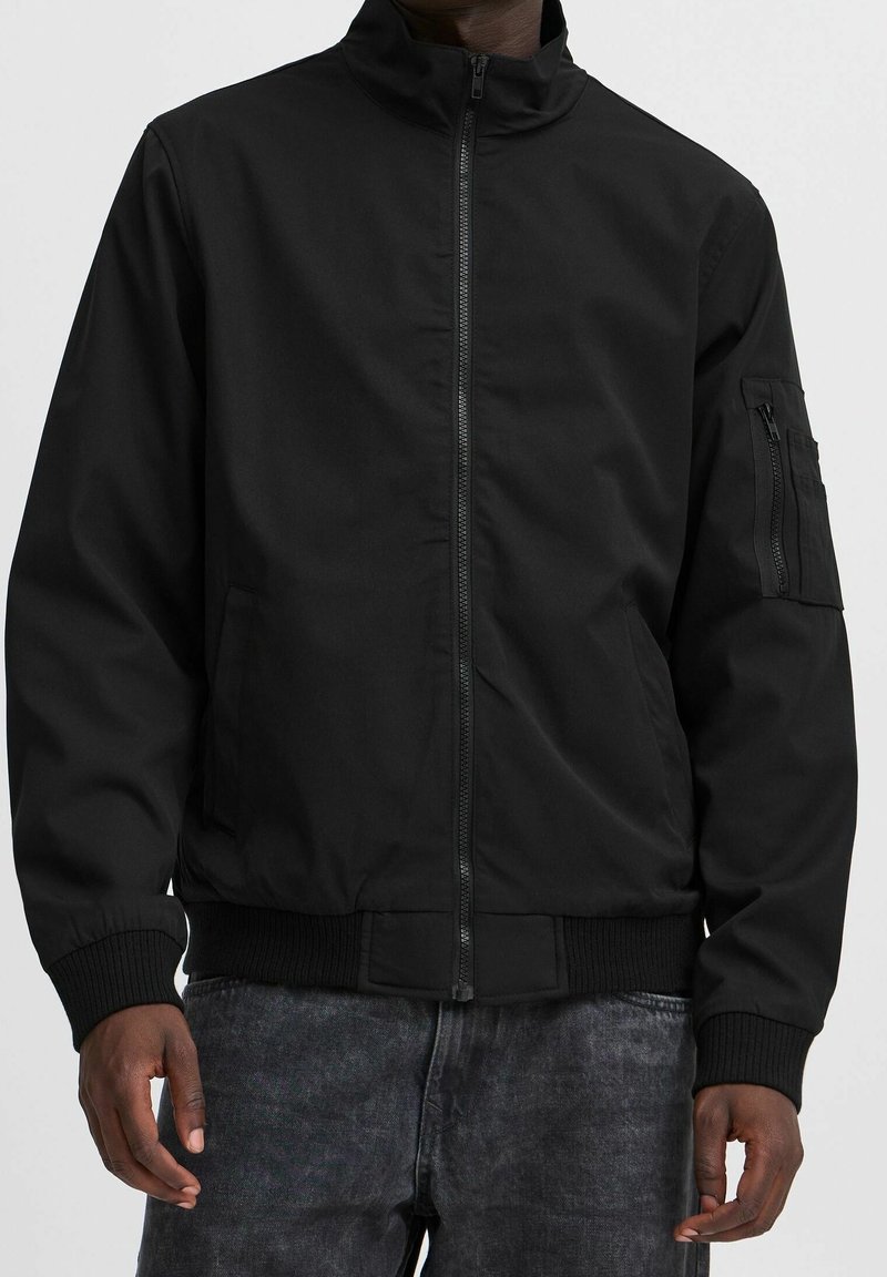 Homme portant une veste bomber noire zippée unie avec des poignets côtelés et une poche zippée sur la manche gauche, associée à un jean foncé.