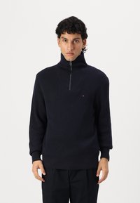 Navy-gestreifter Strickpullover mit hochschließendem Reißverschlusskragen, der über einen quadratischen Ausschnitt und ein kleines Logodetail auf der Brust verfügt.