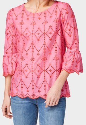 Frau trägt eine rosa Bluse mit roten, gestickten geometrischen Mustern und gewellten Kanten, kombiniert mit blauen Jeans, vor einfachem Hintergrund.