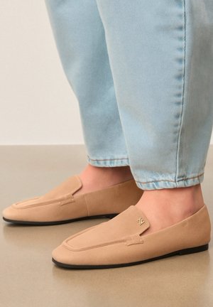 Voeten met beige suède instap loafers met een klein gouden logo, gecombineerd met lichtblauwe opgerolde denim jeans op een gladde vloer.