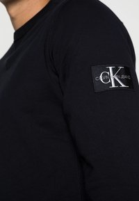 Camicia a maniche lunghe blu navy in cotone, con un tessuto testurizzato e un'etichetta logo rettangolare nera sulla parte superiore del braccio.