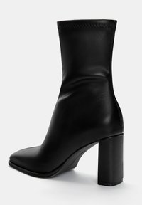 Bottine noire en cuir avec un bout pointu, une texture lisse, un talon haut et épais, et des détails cousus autour de la cheville.