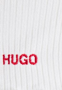 Bílá žebrovaná látka s vyšitým nápisem "HUGO" v červené barvě, obsahující texturované pruhy po celém materiálu.