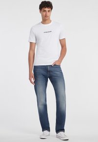 Balta kokvilnas t-krekls ar melnu "GUESS JEANS" logotipu, kombinēts ar zila džinsa biksēm ar izbalējušu mazgāšanu un tradicionālu piecu kabatu dizainu.