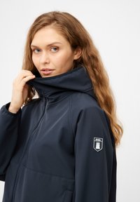 Giacca soft-shell blu navy con colletto alto, zip frontale e patch con logo sulla manica. Presenta una texture liscia e un design minimale.