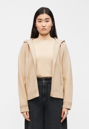 Femme aux longs cheveux foncés portant un sweat à capuche beige zippé sur un haut beige et un jean noir taille haute, debout devant un fond uni.