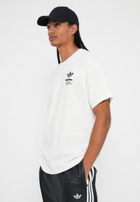 White cotton t-shirt with a crew neck, featuring a black Adidas logo on the chest. Model wears black cap and matching black track pants.