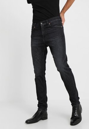 Homme portant un jean noir slim et des bottines en cuir noir, debout devant un fond gris clair uni.