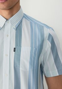 Kratkoslečen srajc v svetlo modrih in belih črtah, ki ima žep na prsih ter klasičen ovratnik z diskretno podrobnostjo logotipa.