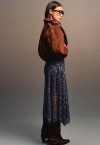 Brun ruskinds oversized jakke, blåternet lang nederdel og brune støvler til knæet. Solbriller med et dristigt, vinklet design. Sideprofilbillede.