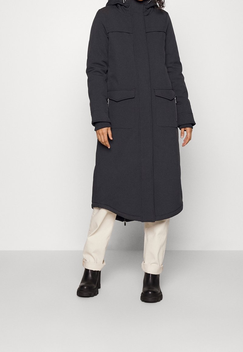 Manteau matelassé long gris foncé avec capuche, doté de deux poches avant, d'une fermeture éclair et d'un ourlet droit. Porté sur un pantalon clair.