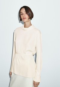 Haut créme à manches longues avec col drapé, taille cintrée, texture lisse et coupe ajustée. Design simple et minimal sans motifs visibles.