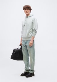 Νέος άνδρας με σγουρά μαλλιά φοράει μια ανοιχτό γκρι κουκούλα, ξεθωριασμένο μπλε τζιν, μαύρα παπούτσια και κρατάει μια μαύρη δερμάτινη σακούλα duffle.