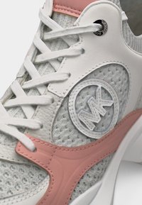 Κοντινό πλάνο λευκού και ροζ αθλητικού παπουτσιού με ύφασμα από δίχτυ, δερμάτινες επενδύσεις, λευκά κορδόνια και κυκλικό λογότυπο MK στην πλαϊνή πλευρά.