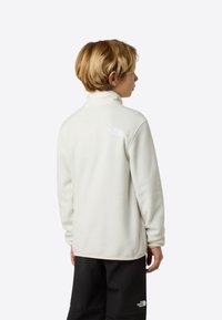 Biały polarowy pulower z wysokim kołnierzem, z małym logo na górnej części pleców. Miękka tekstura, w zestawieniu z czarnymi spodnimi elementami.