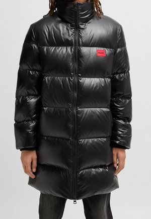 Personne portant un long manteau doudoune noir brillant avec une fermeture éclair à l'avant et un petit logo rouge Hugo sur la poitrine.