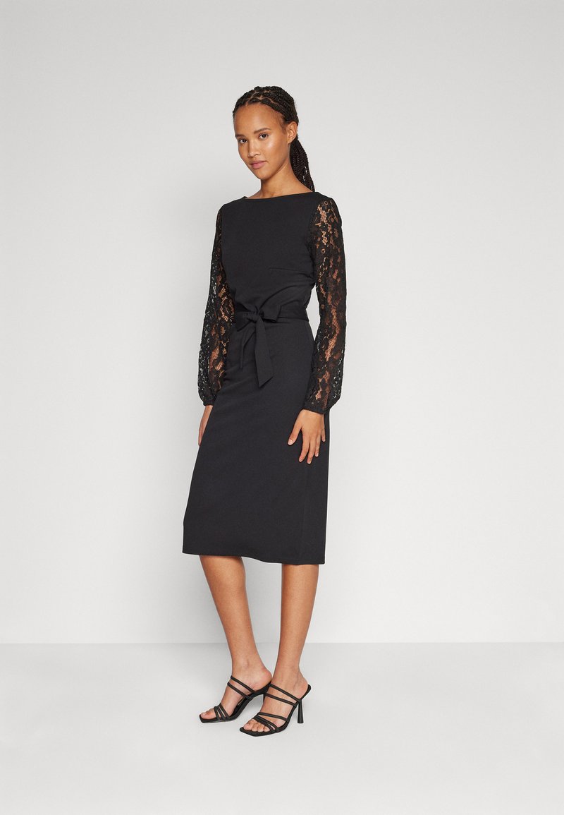 Robe noire avec des manches longues en dentelle, corps solide, détail de ceinture à nouer, longueur au genou, associée à des escarpins noirs à lanières sur un fond uni.