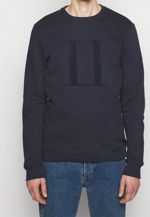 Mężczyzna w granatowej bluzie z delikatnym pionowym podwójnym wzorem, w połączeniu z niebieskimi dżinsami, na jednolitym tle.