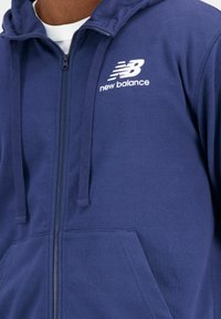 Marineblauwe zip-up hoodie van zachte stof, met een wit "New Balance" logo op de borst, koordjes en een voorzak.