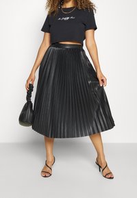 Jupe plissée noire avec une finition brillante, associée à un t-shirt noir ajusté et court. Accessoirisée avec un petit sac noir et des sandales noires.