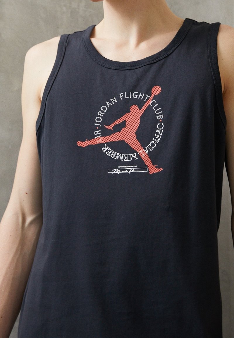 Homme portant un débardeur noir avec une silhouette rouge d’un joueur de basket-ball et un texte circulaire « Jordan Flight Club Official Member ».