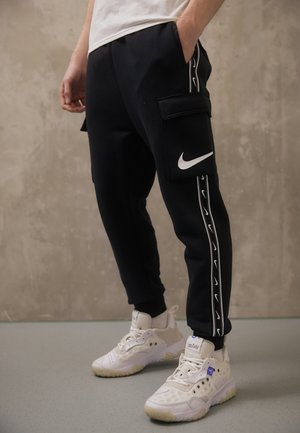 Zwarte joggingbroek met een cargozak, wit Nike-logo en zijkantape-detail. Gecombineerd met witte sneakers met doorschijnende zolen.