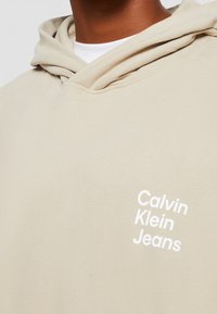 Béžová mikina s kapucí vyrobená z bavlněné směsi. Bílé logo "Calvin Klein Jeans" tištěné na levé části hrudi. Měkká textura.