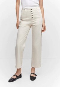 Pantalones crema de cintura alta con un cierre frontal de botones, diseño de pierna recta y sutilezas en las costuras. Combinados con zapatos planos negros.
