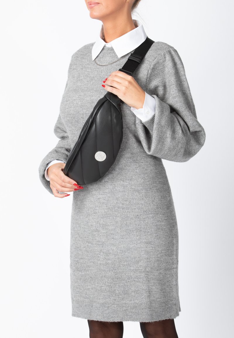 Borsa a tracolla in sintetico nero con design trapuntato e un accento circolare in argento. Il modello indossa un vestito maglione grigio e una camicia bianca con colletto.