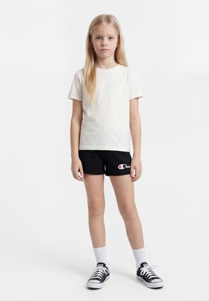 Enfant blond debout, portant un t-shirt blanc, un short noir avec logo, des chaussettes blanches et des baskets noires, sur un fond blanc uni.