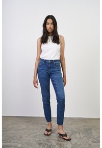 Top blanco sin mangas combinado con jeans de mezclilla azul ajustados, con cintura alta y un ligero deshilachado en los tobillos, complementado con chanclas marrones.