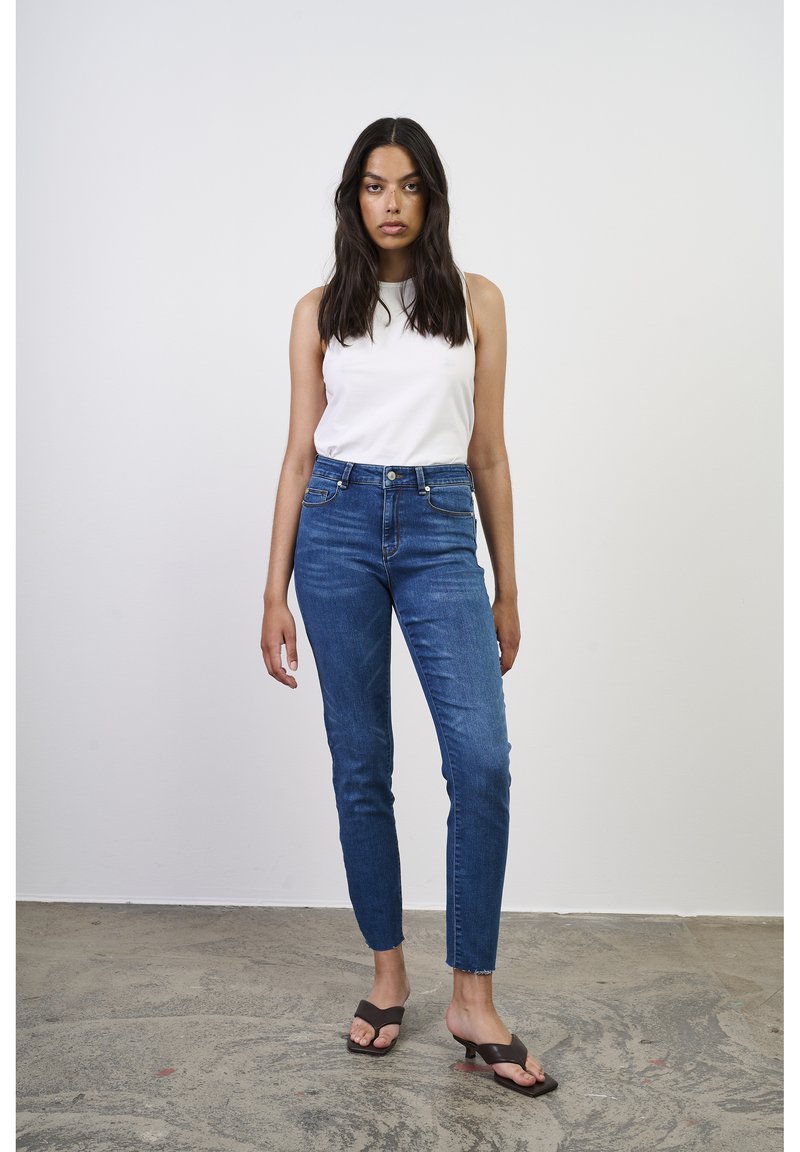 Top blanco sin mangas combinado con jeans de mezclilla azul ajustados, con cintura alta y un ligero deshilachado en los tobillos, complementado con chanclas marrones.