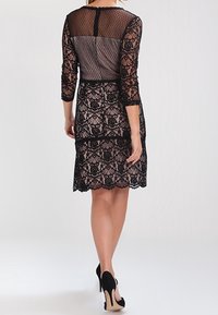 Femme portant une robe noire en dentelle arrivant aux genoux avec des manches transparentes et un dos dénudé, s'éloignant, assortie à des chaussures à talons hauts noires.