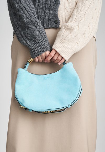 Light blue suede handbag with a curved shape, gold hardware accents, and a top handle, held by two hands against a neutral background.